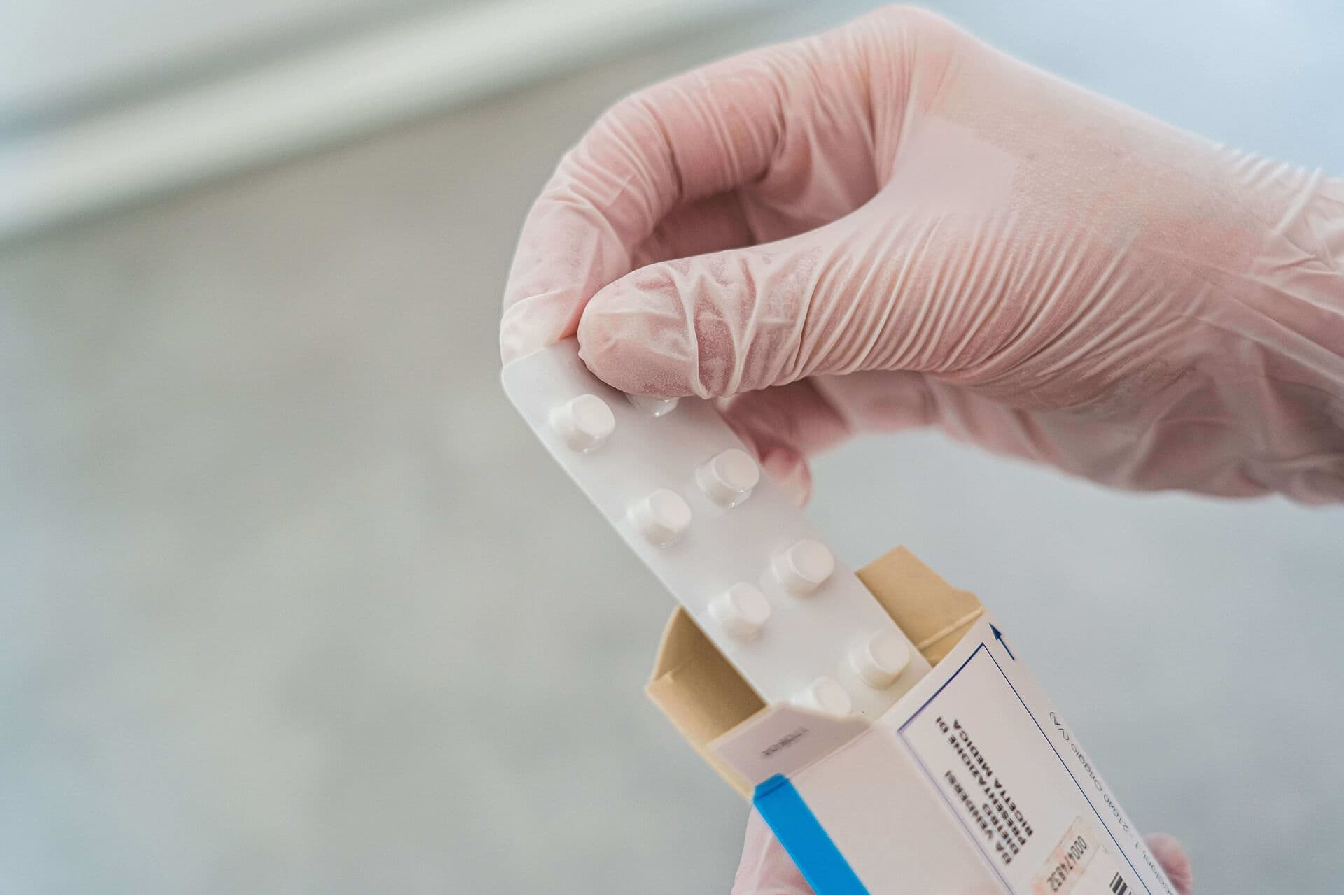 A gloved hand lifting a blister strip of tablets out of its printed carton